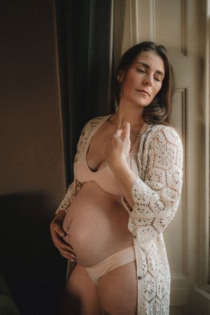 Intimate maternity portraits featuring a woman in a lace robe posing near natural window light.