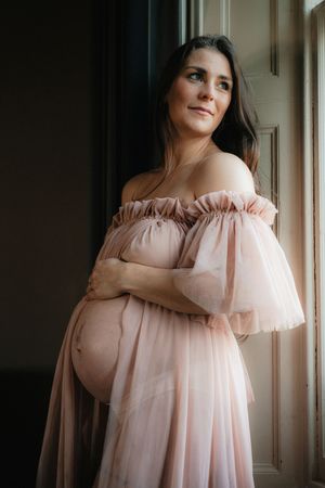 A woman in a soft pink tulle dress poses in a series of romantic indoor portraits near a window.