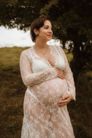 Maternity photo session featuring a delicate white lace dress against a natural outdoor backdrop.