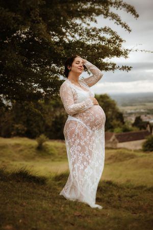 Artistic maternity portraits in white lace dress against rolling hills and moody sky.