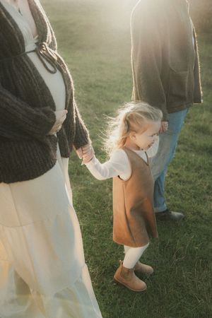 A small child in a brown dress and boots holds hands with adults while standing on grass in golden sunlight.