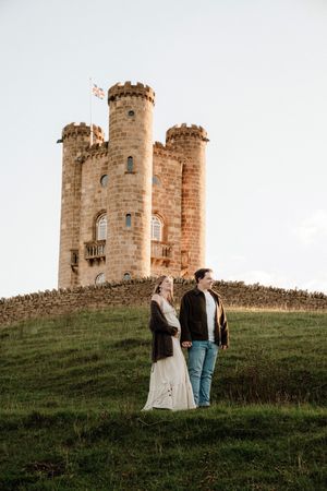 Medieval stone castle tower stands atop grassy hill during sunset with two figures silhouetted against warm evening light.