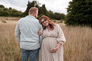 A couple stands together in a golden field during sunset, with the expectant mother wearing a flowing cream dress.
