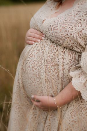 Close-up shots of a pregnant belly draped in delicate cream lace dress showing the curve of maternity in natural light.