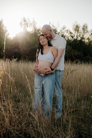 A couple embraces in a sunlit field of tall grass during a maternity photoshoot at golden hour.
