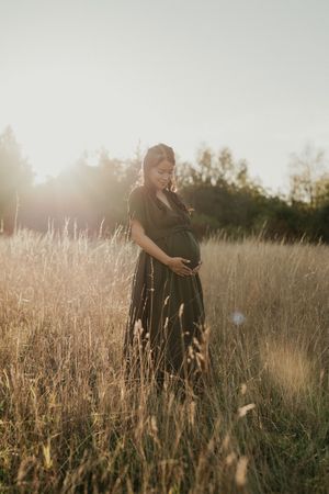 A silhouetted figure in a flowing dress stands among tall grass during a sunset maternity photo session.