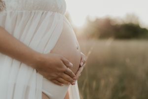 A person in a flowing white dress gently cradles their pregnant belly during a golden hour maternity photoshoot.