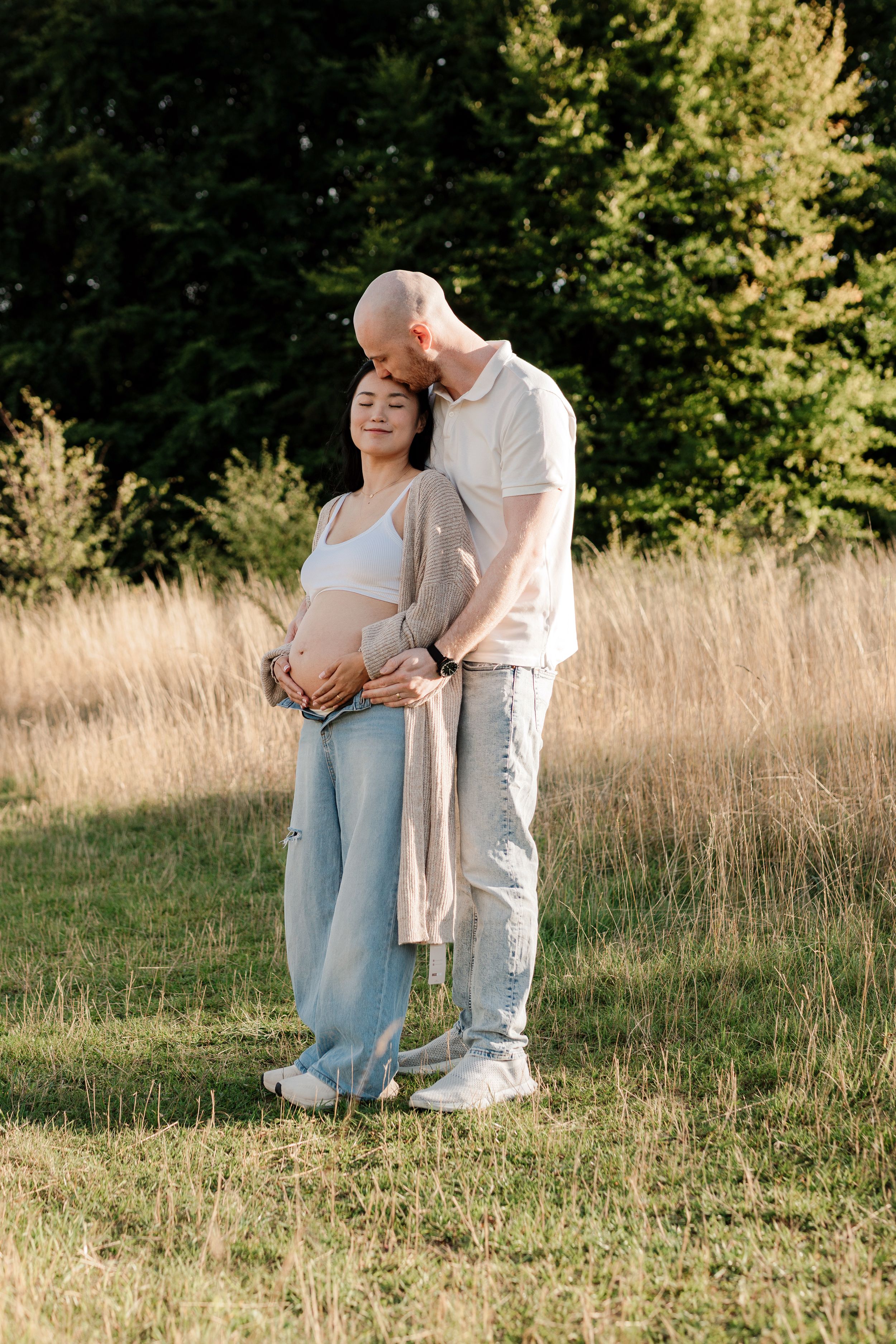 Couple embraces in golden field with evergreen trees during sunset maternity photoshoot.