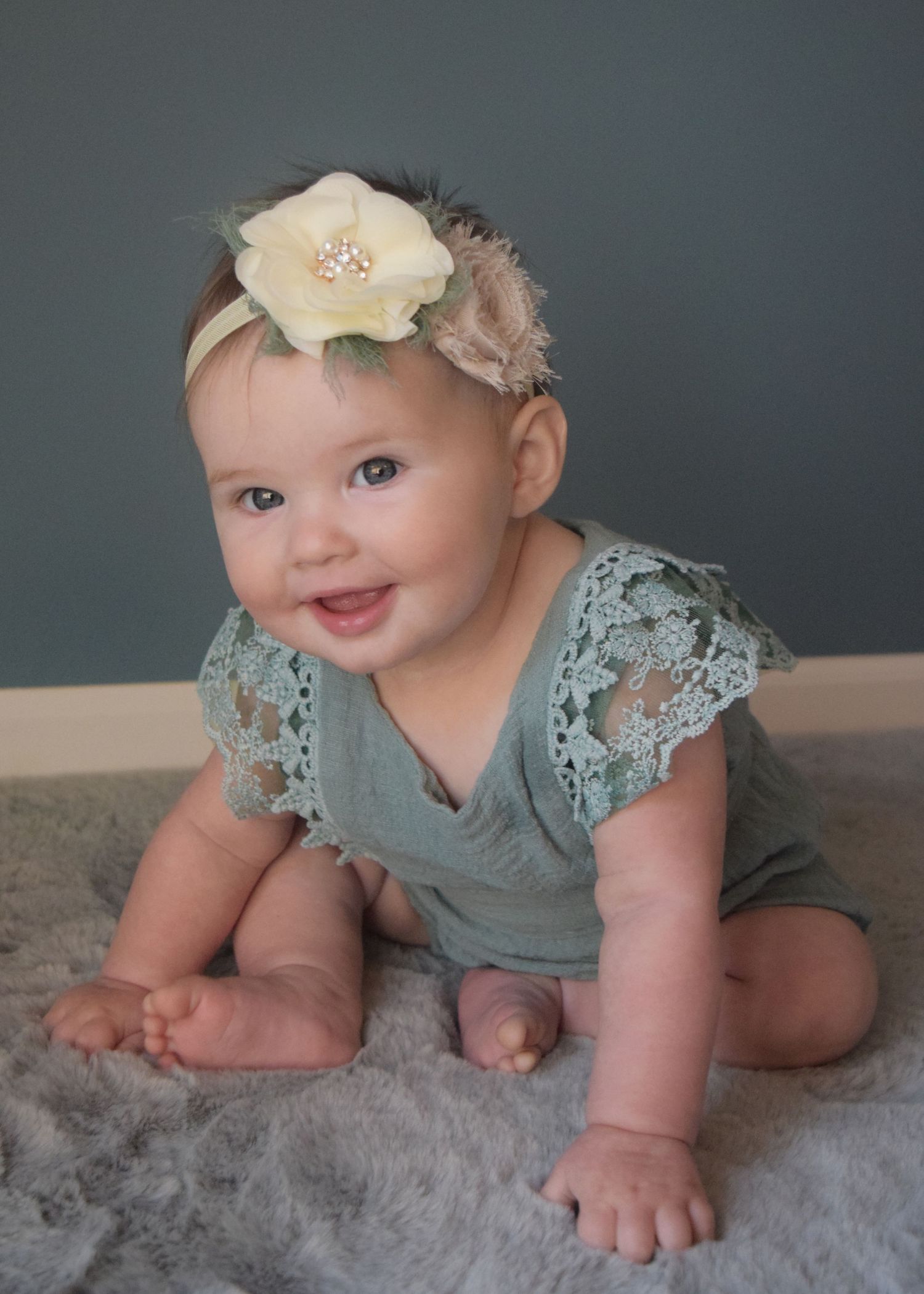 Baby in sage green lace outfit sitting on plush gray carpet wearing floral headband.
