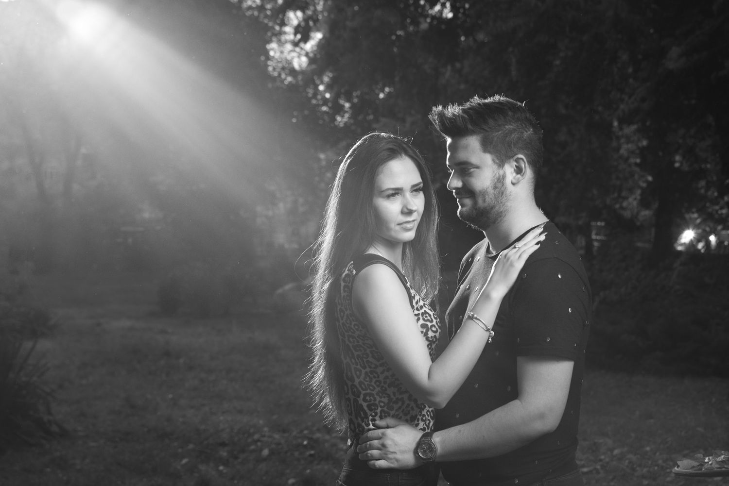 Couple shares intimate moment in misty forest with dramatic lighting and shadows.