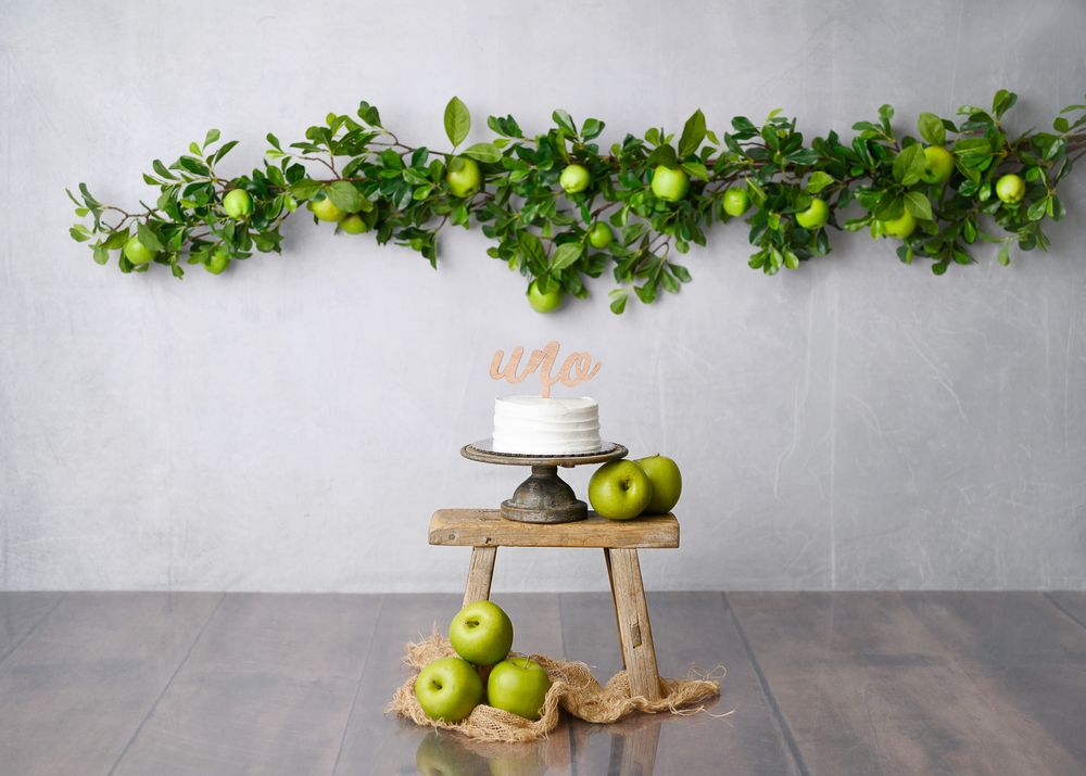 Simple white cake on rustic wooden stand with green apple decor and leafy garland above.