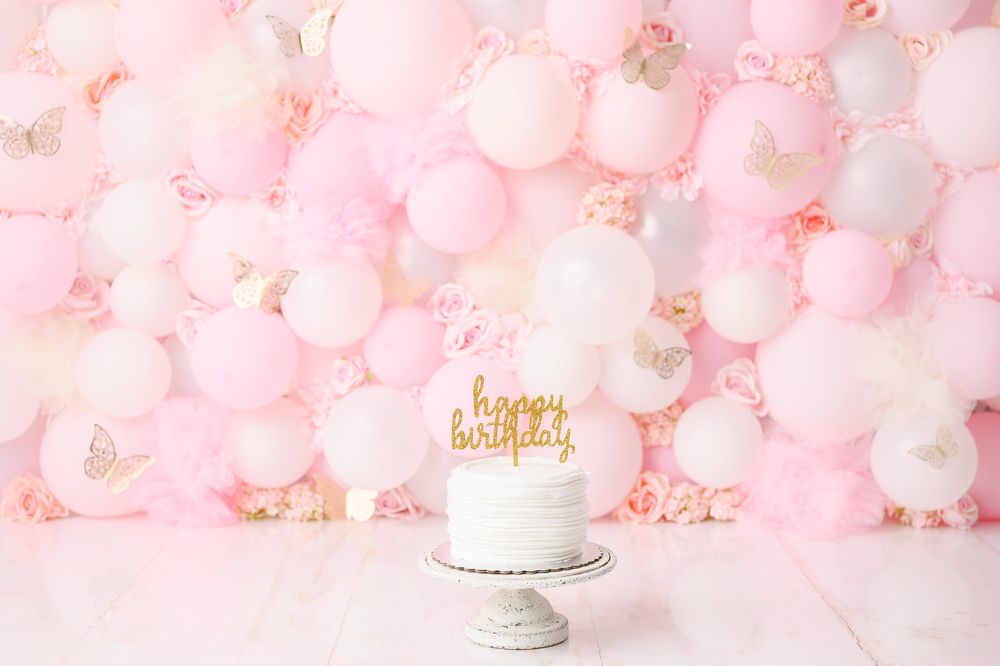 White birthday cake against pink balloon wall backdrop with gold 'happy birthday' cake topper.