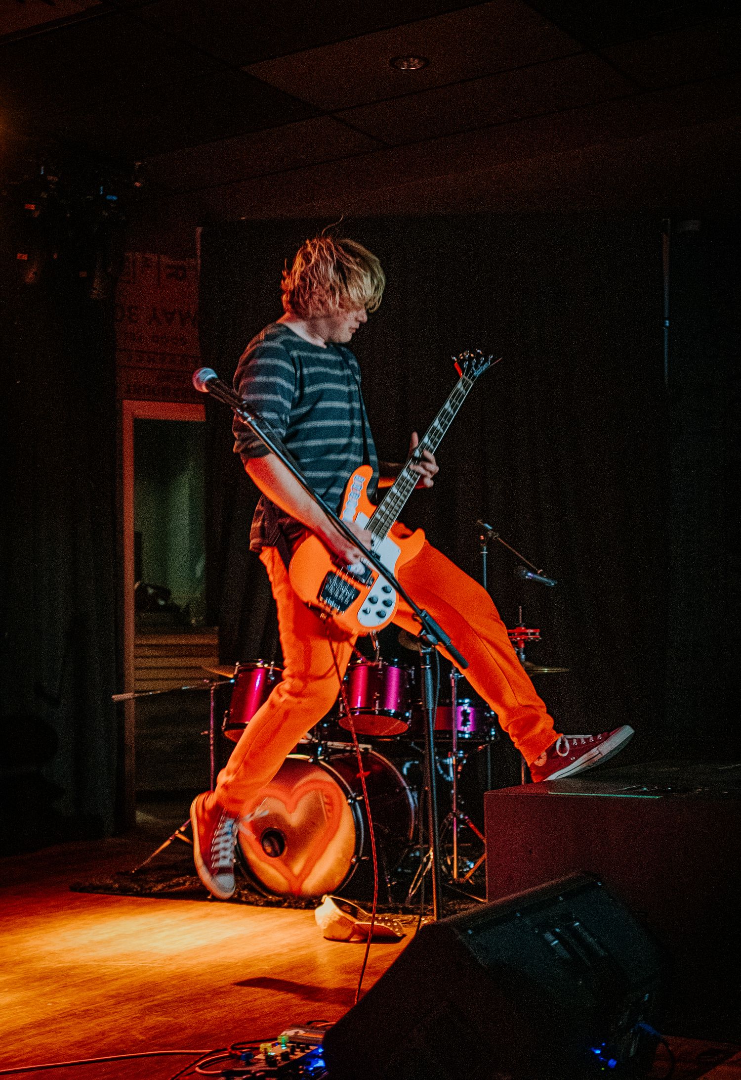 Guitarist in striped shirt and orange pants performs on brightly lit stage with drum kit visible.