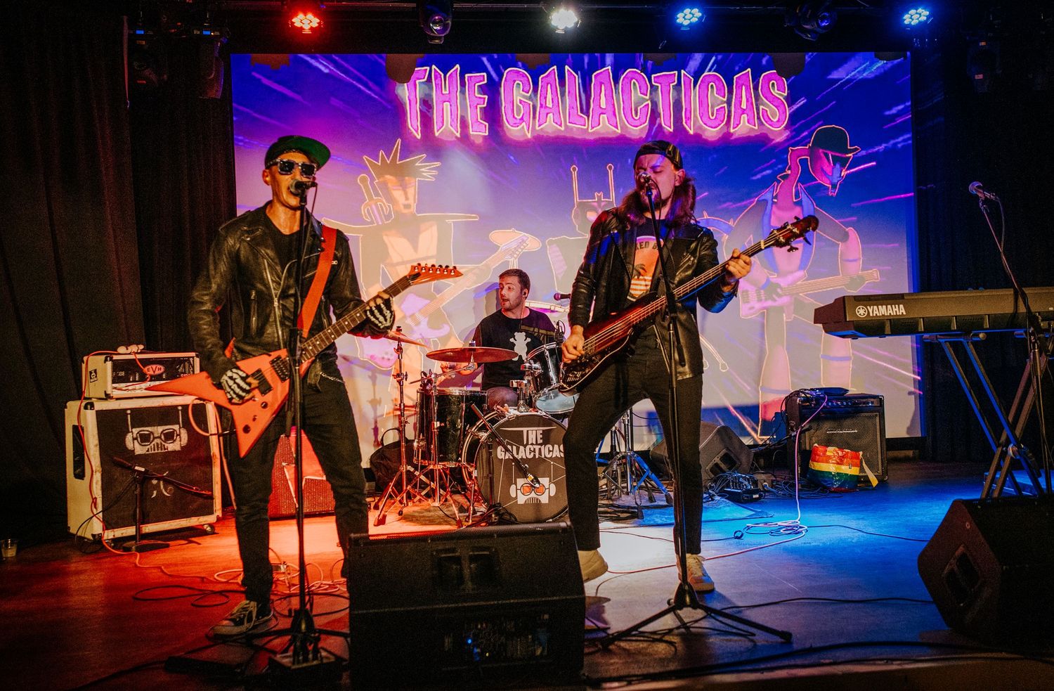 Rock band performs on stage with illuminated band name 'The Galactics' displayed behind them.
