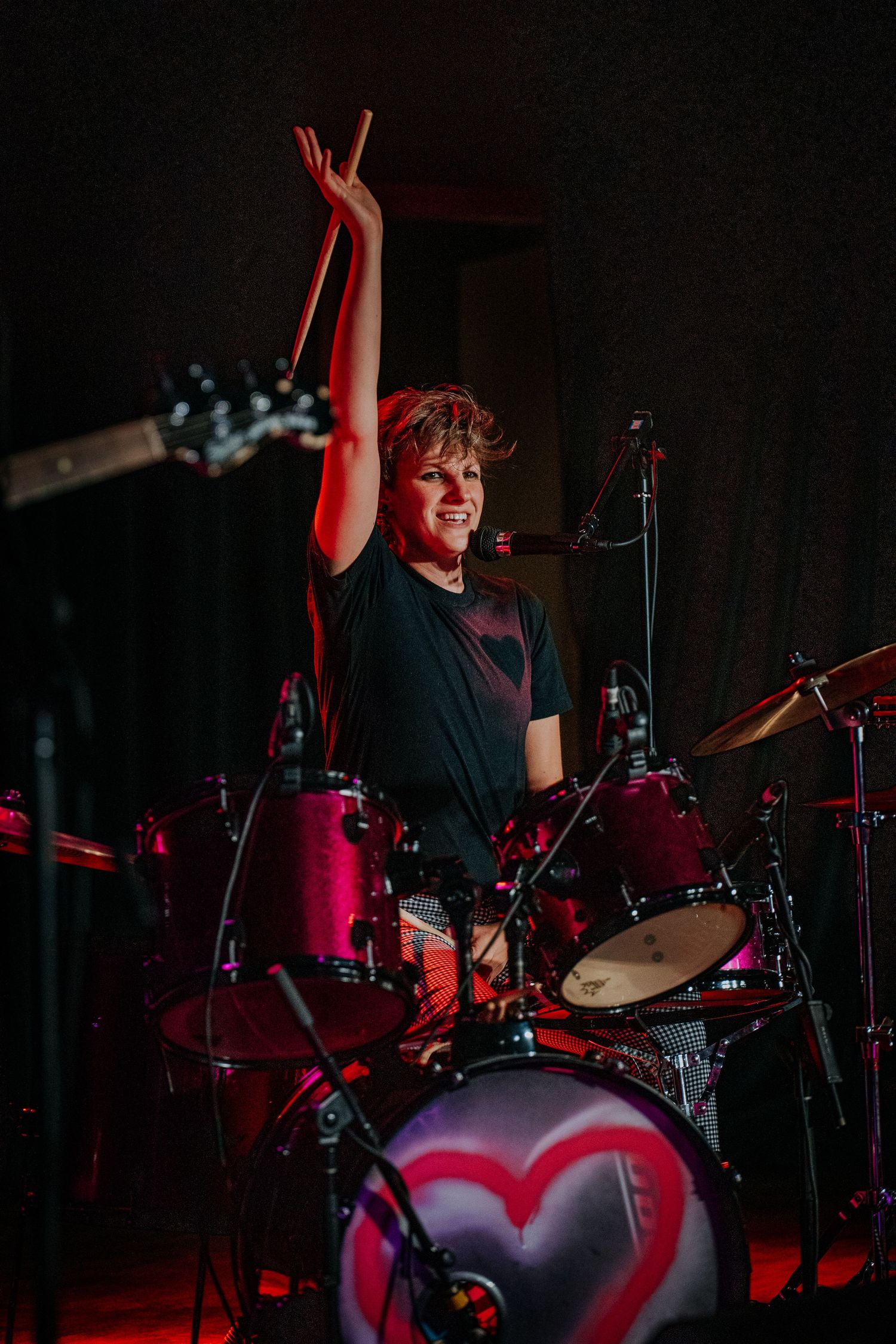 Drummer raises arm triumphantly behind drum kit during energetic performance under red stage lights.