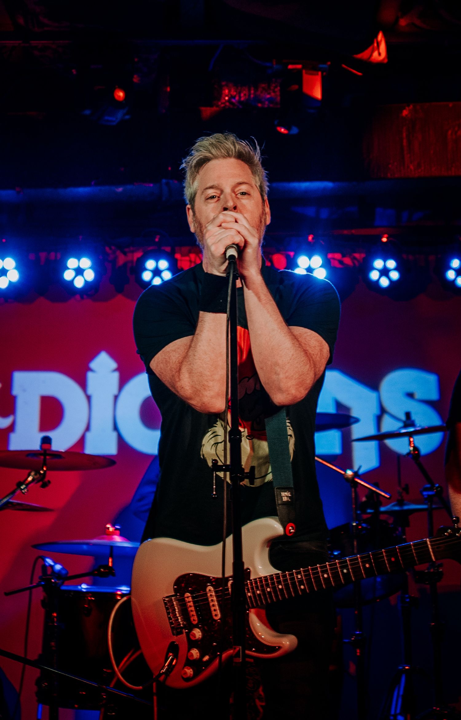 Musician singing into microphone on stage with blue LED lights and band logo in background.