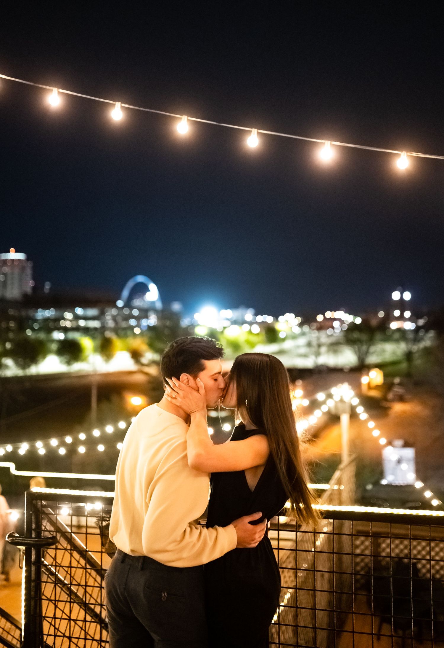 A romantic nighttime embrace under string lights with a city skyline glowing in the background.
