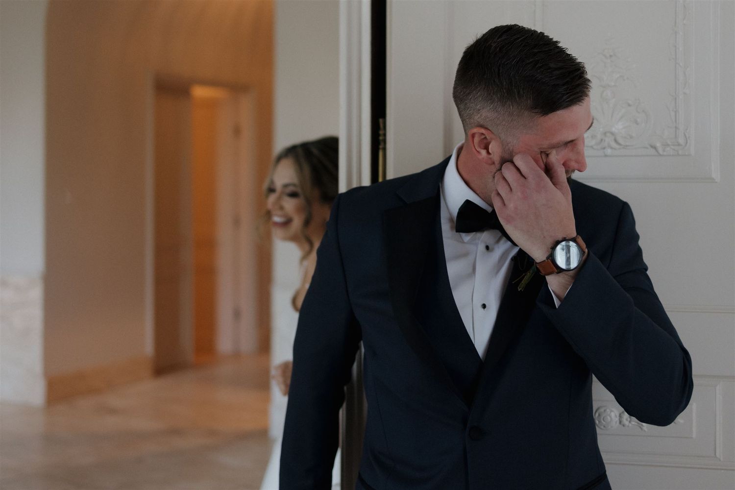 An emotional first look moment at a wedding as the groom wipes his eyes while wearing a black tuxedo.