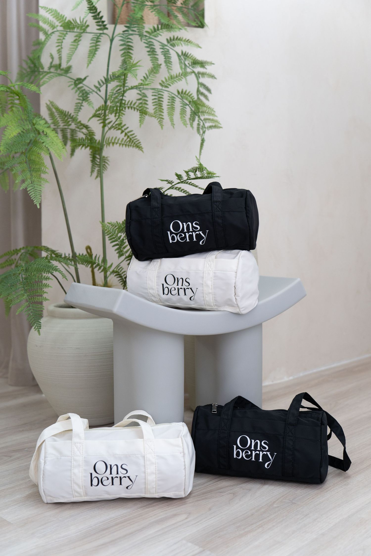 Display of black and white Ons berry duffle bags arranged on modern gray table with fern plant.