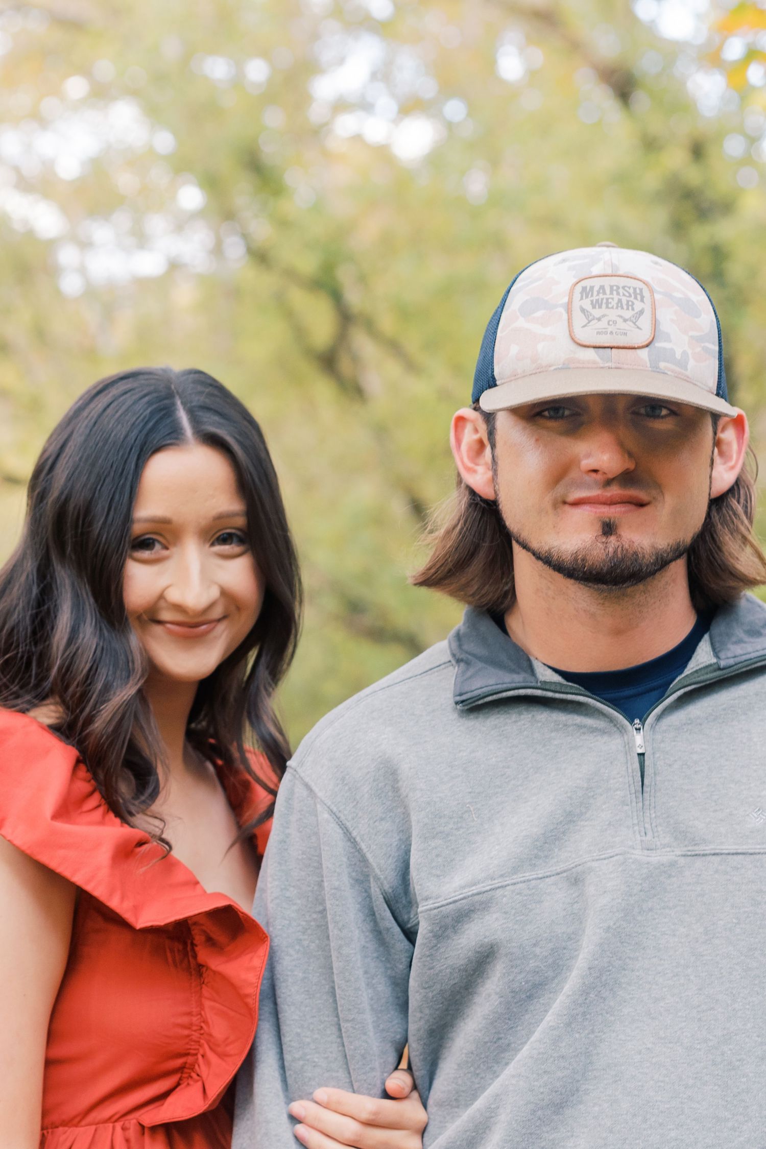 A couple poses outdoors during autumn wearing casual attire against a blurred natural background.