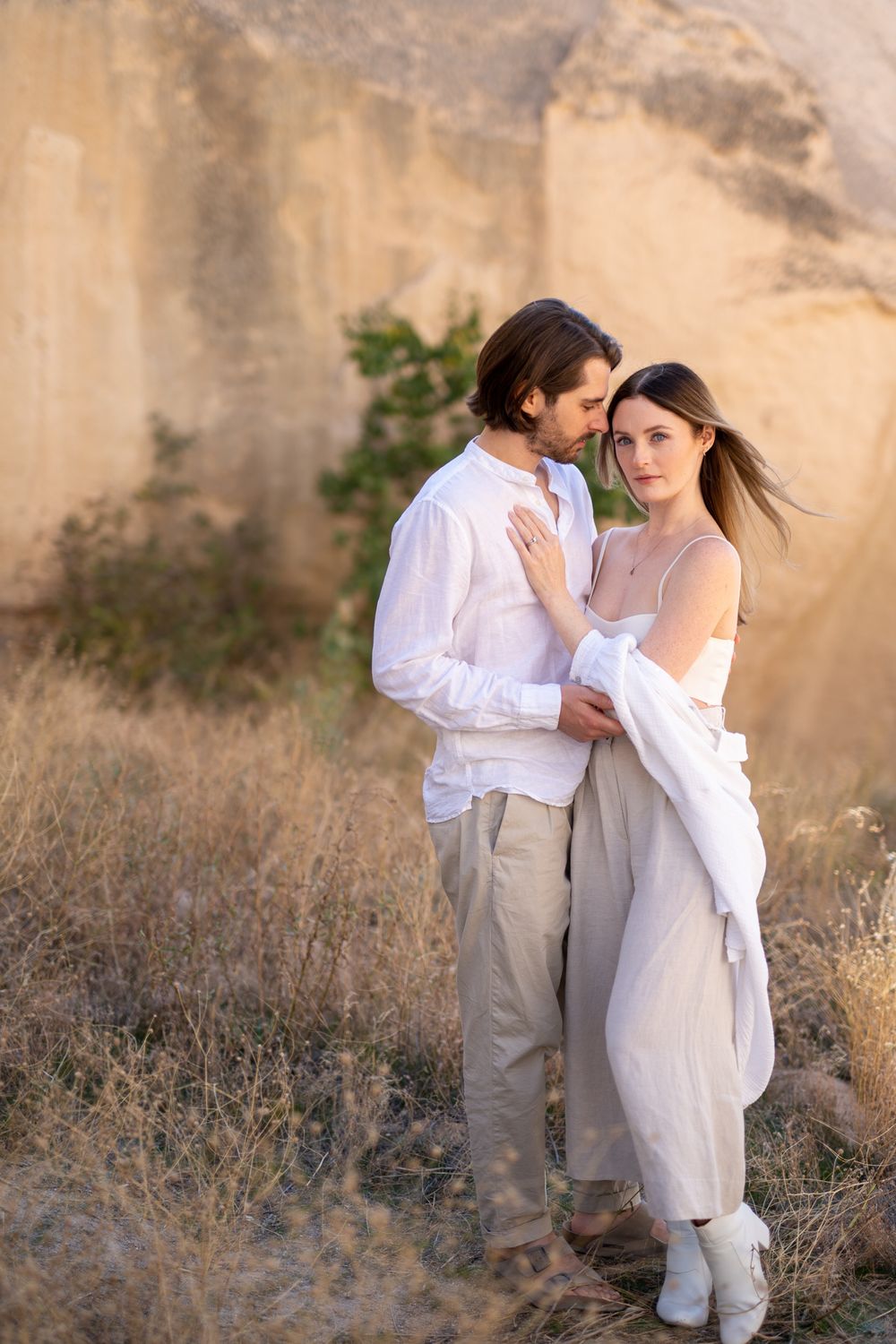 Dreamy couple's portrait in ethereal white outfits against textured stone wall at sunset.