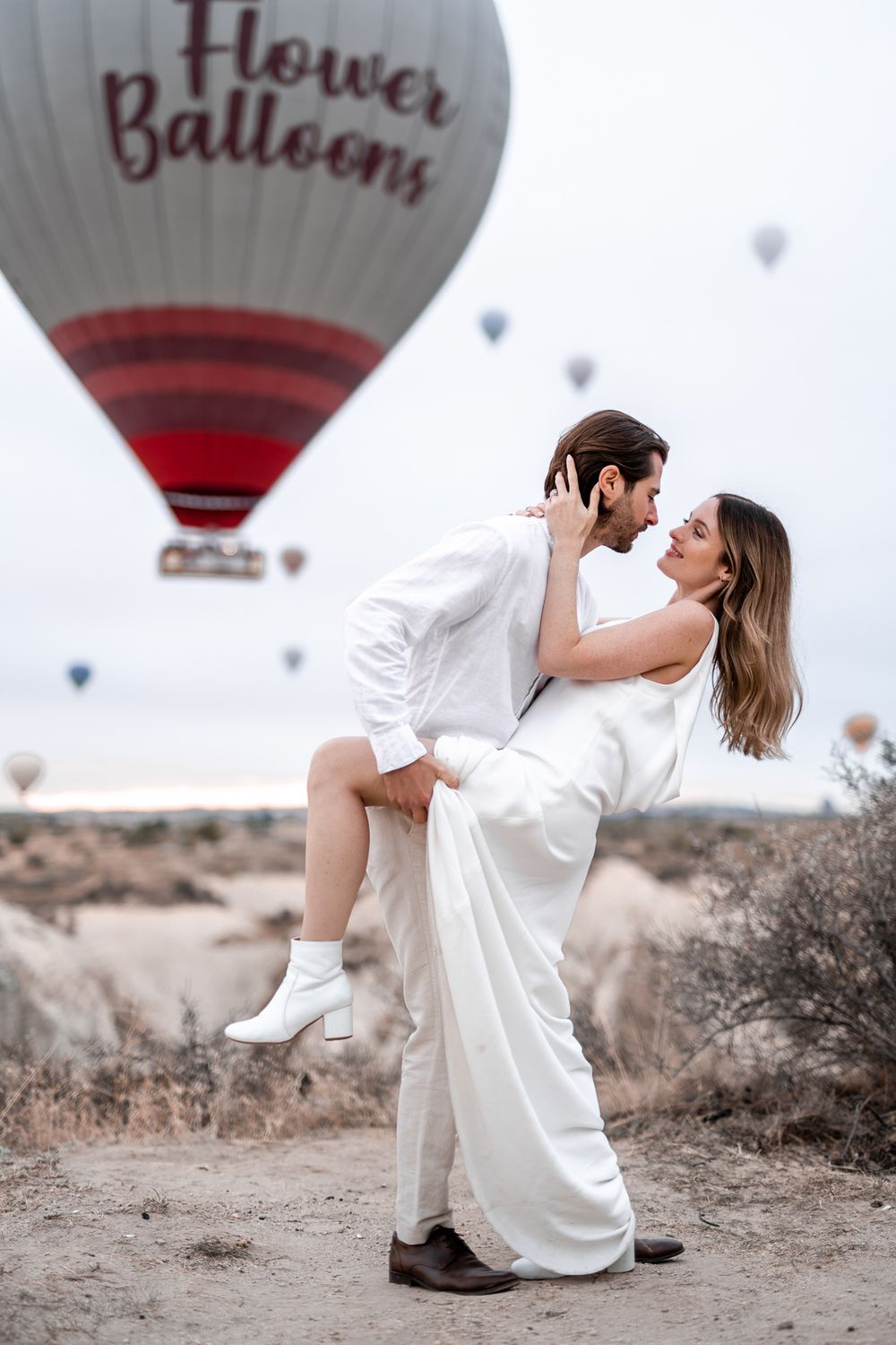 A romantic dip pose in front of a red and white hot air balloon in a desert landscape.