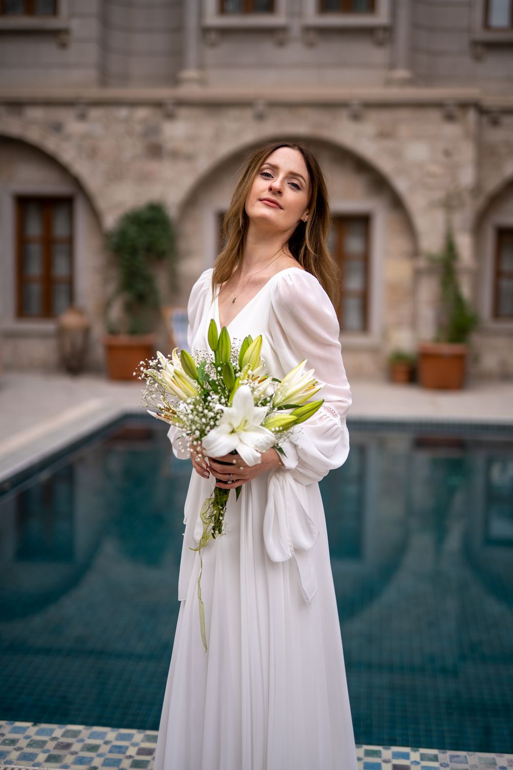 An elegant white dress against a pool backdrop with a beautiful white floral bouquet.