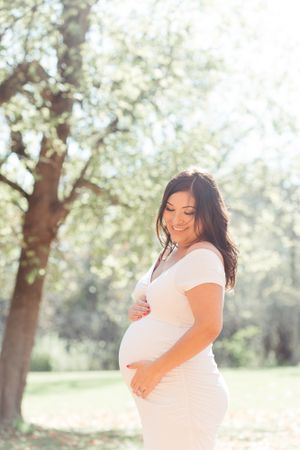 A woman in a white dress poses outdoors during a maternity photoshoot.