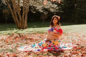 Expectant mother in vibrant floral dress sits among fallen autumn leaves during maternity photoshoot.