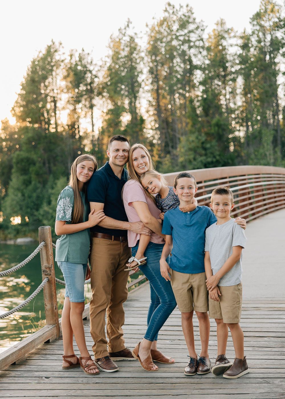 Family portrait on a wooden bridge with pine trees in the background during sunset.