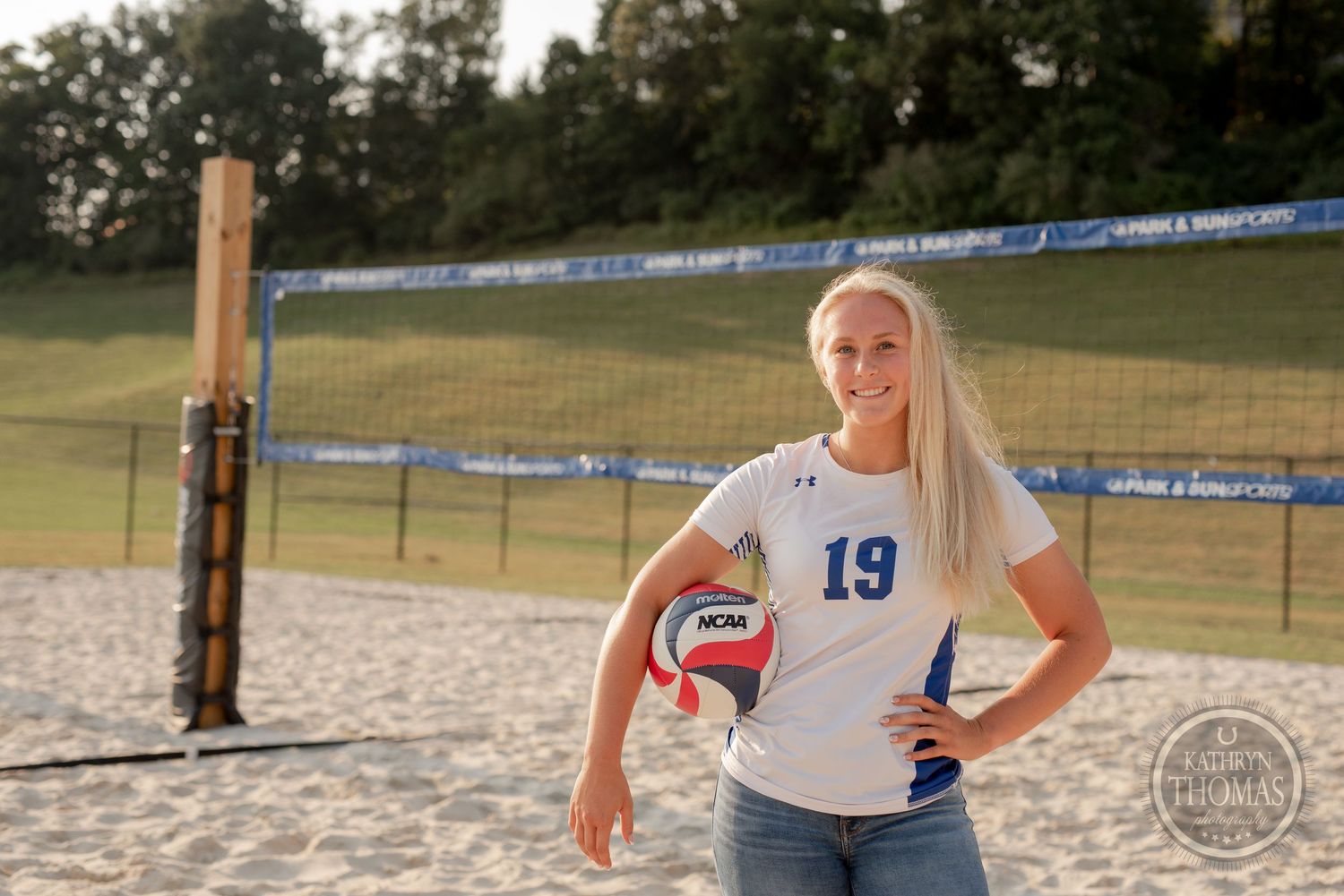 A volleyball player holds a ball while posing on a sandy court wearing jersey number 19.