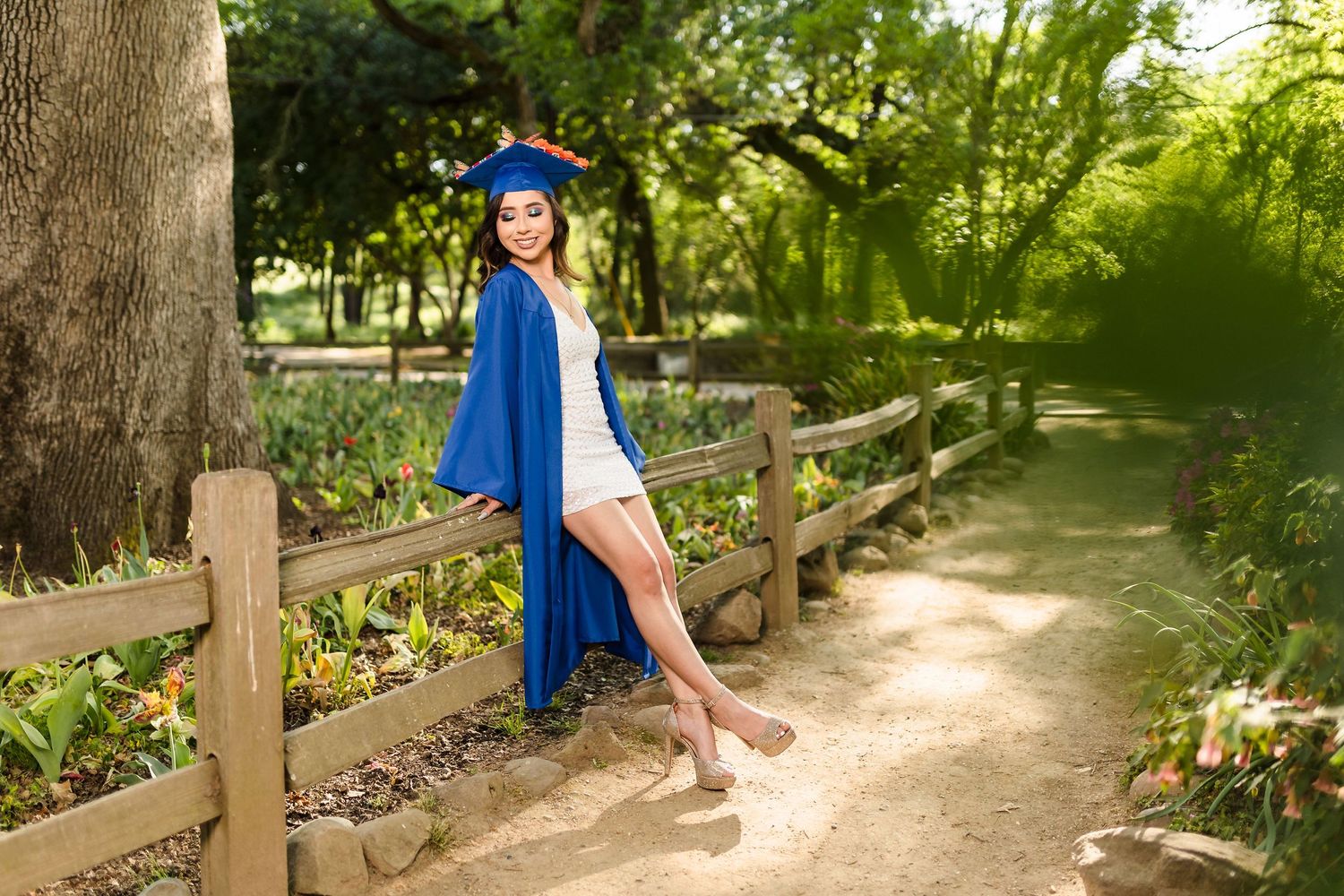 A dancer in blue poses gracefully on a wooden fence along a tree-lined park path.