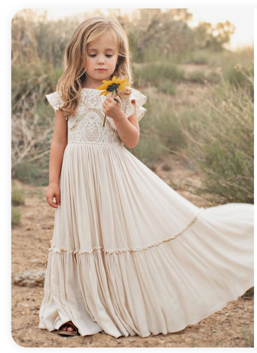 A small girl in a flowing cream-colored lace dress holding a yellow flower in a rustic outdoor setting.