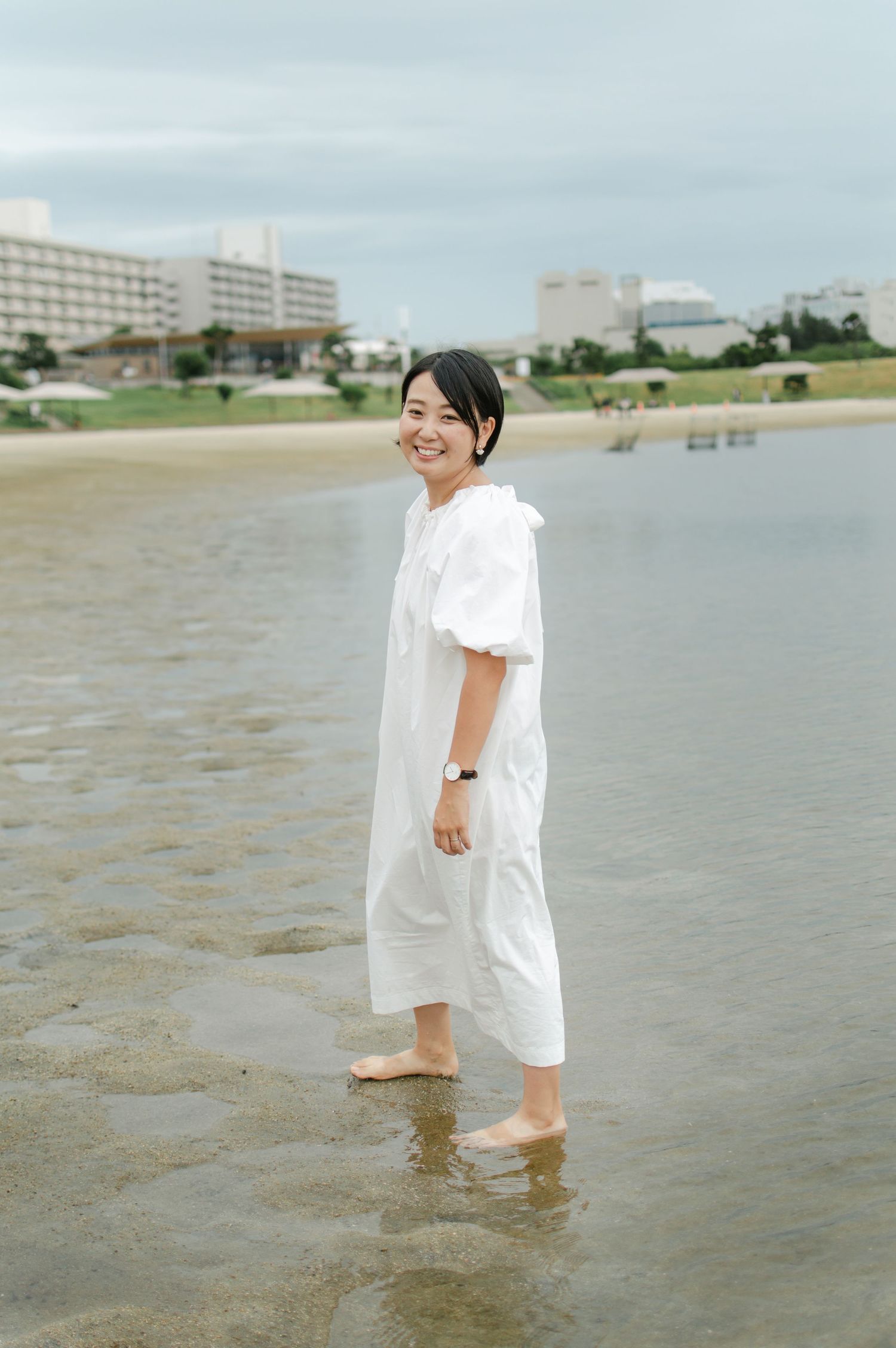 A cheerful woman in a white dress smiles while standing barefoot in shallow beach water.
