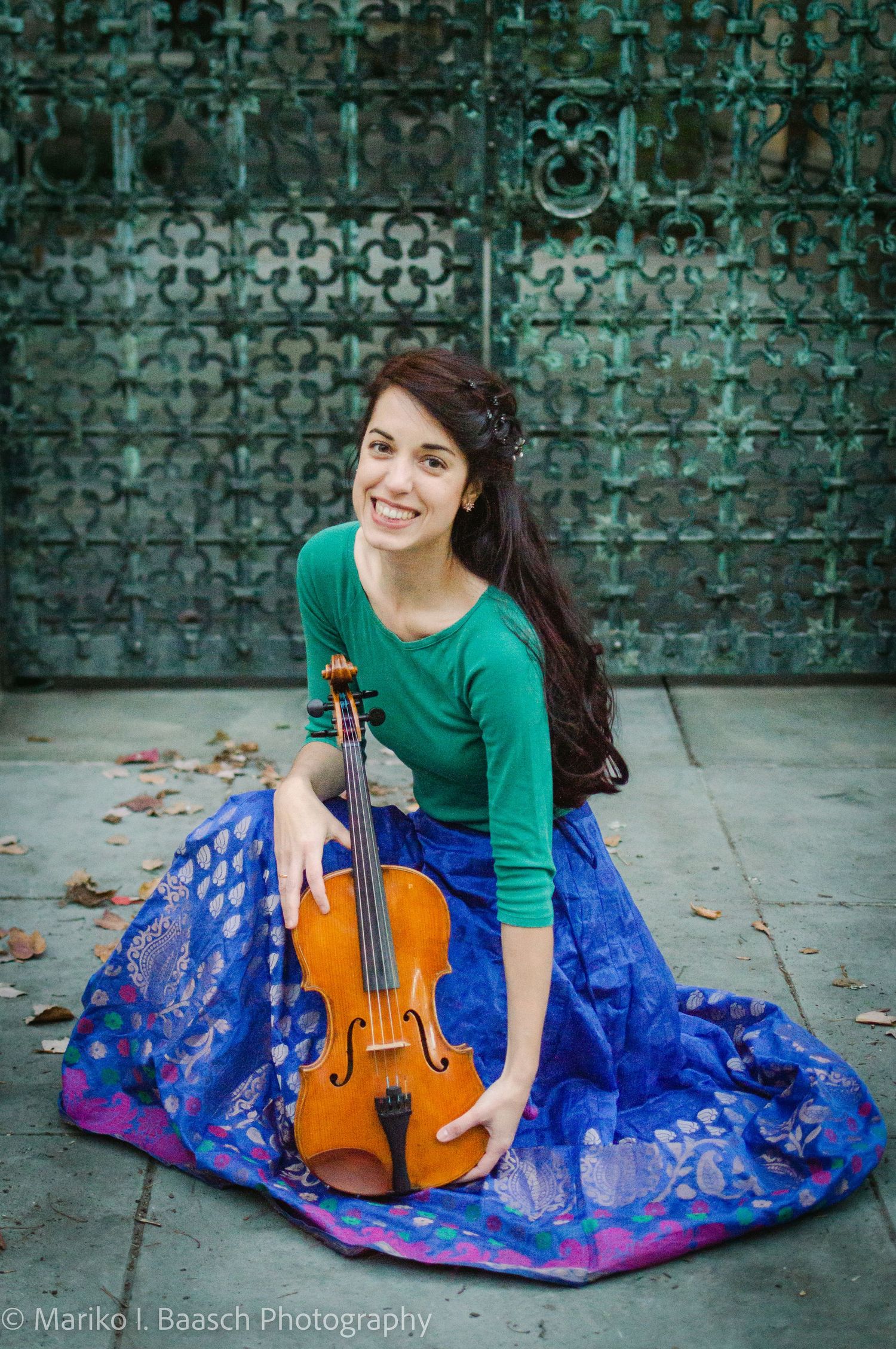 Smiling violinist in blue lehenga sitting on ground holding violin before decorative green gate.
