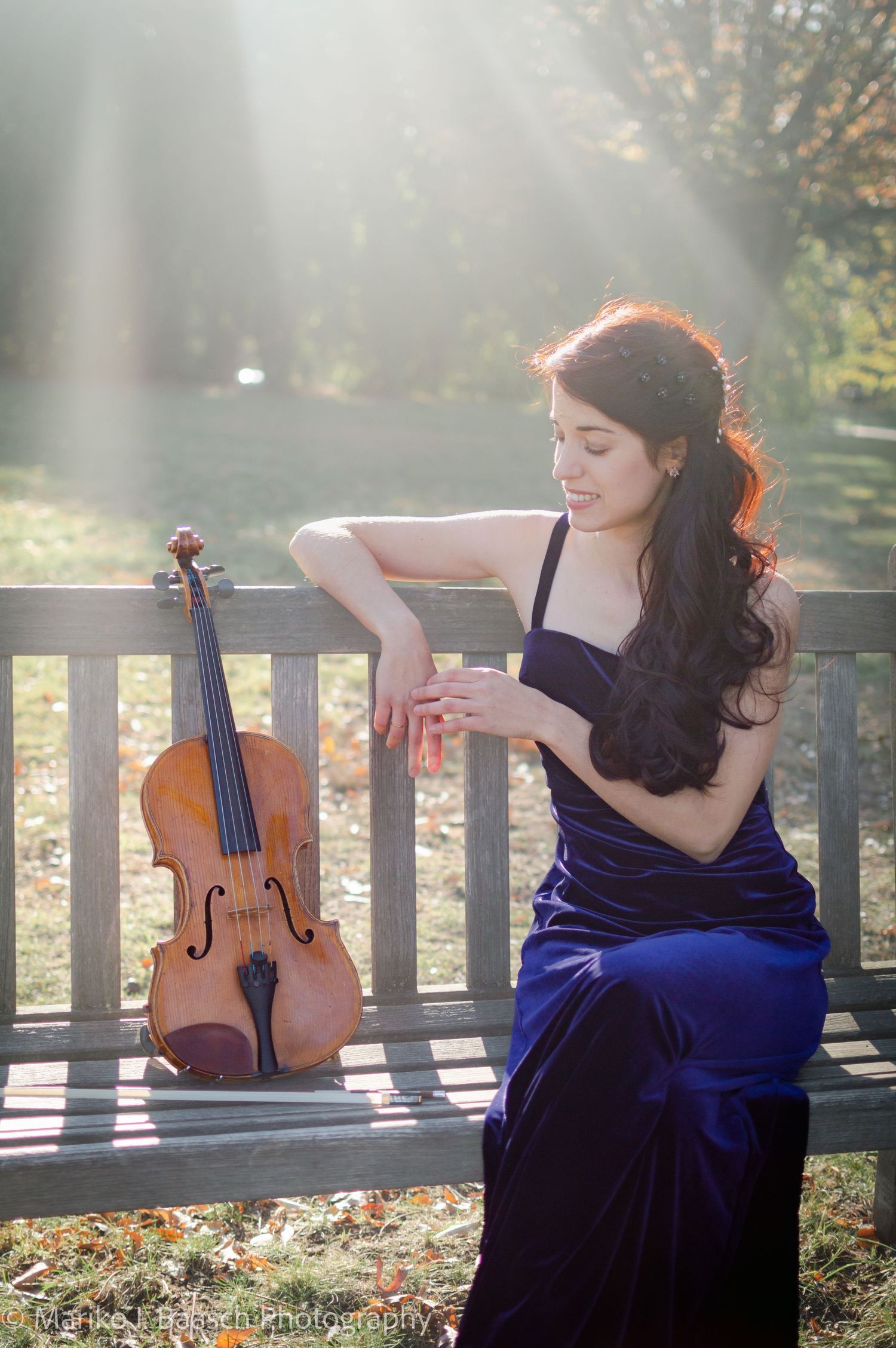 A violinist in a blue velvet gown sits on a bench with her violin in golden sunlight.