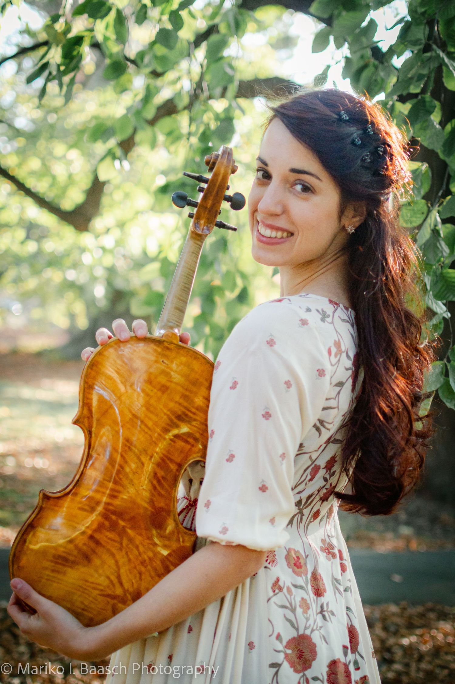 A smiling violinist holds her instrument while posing under green trees in a floral dress.