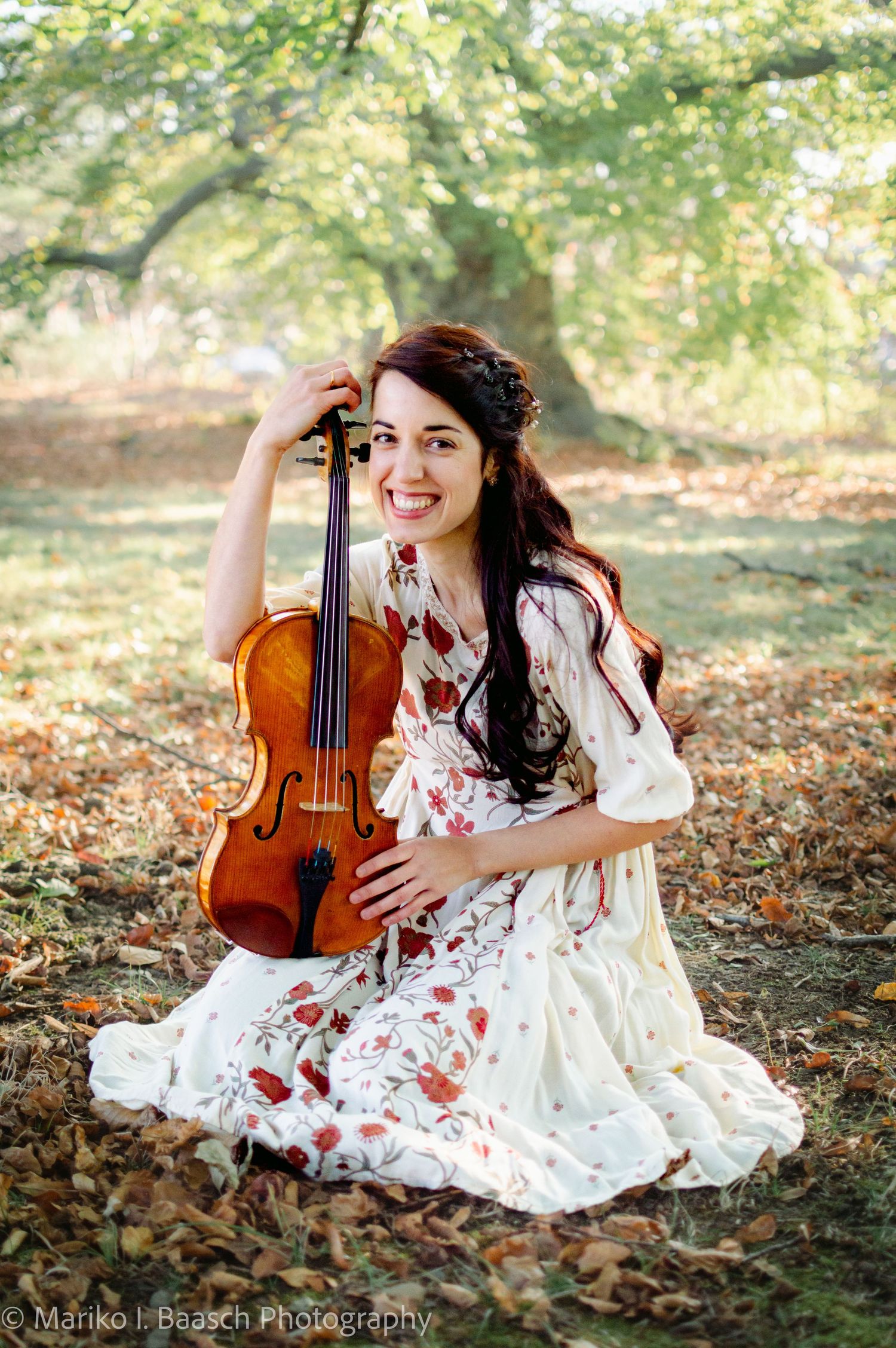A smiling violinist in a floral dress sits among autumn leaves holding her violin.