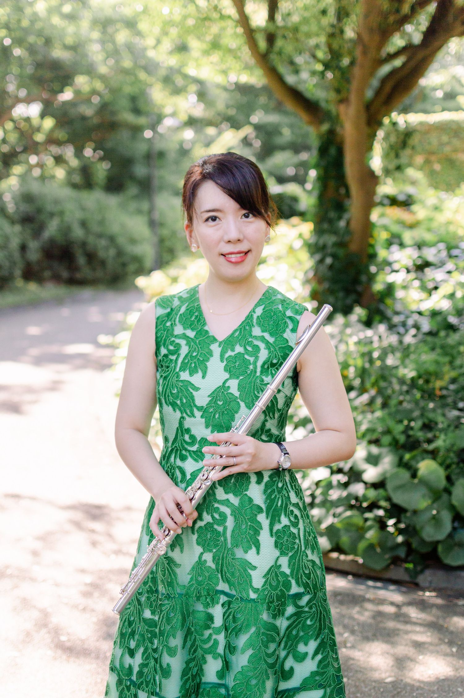 A woman in a green lace dress holds a silver flute while posing in a garden path.