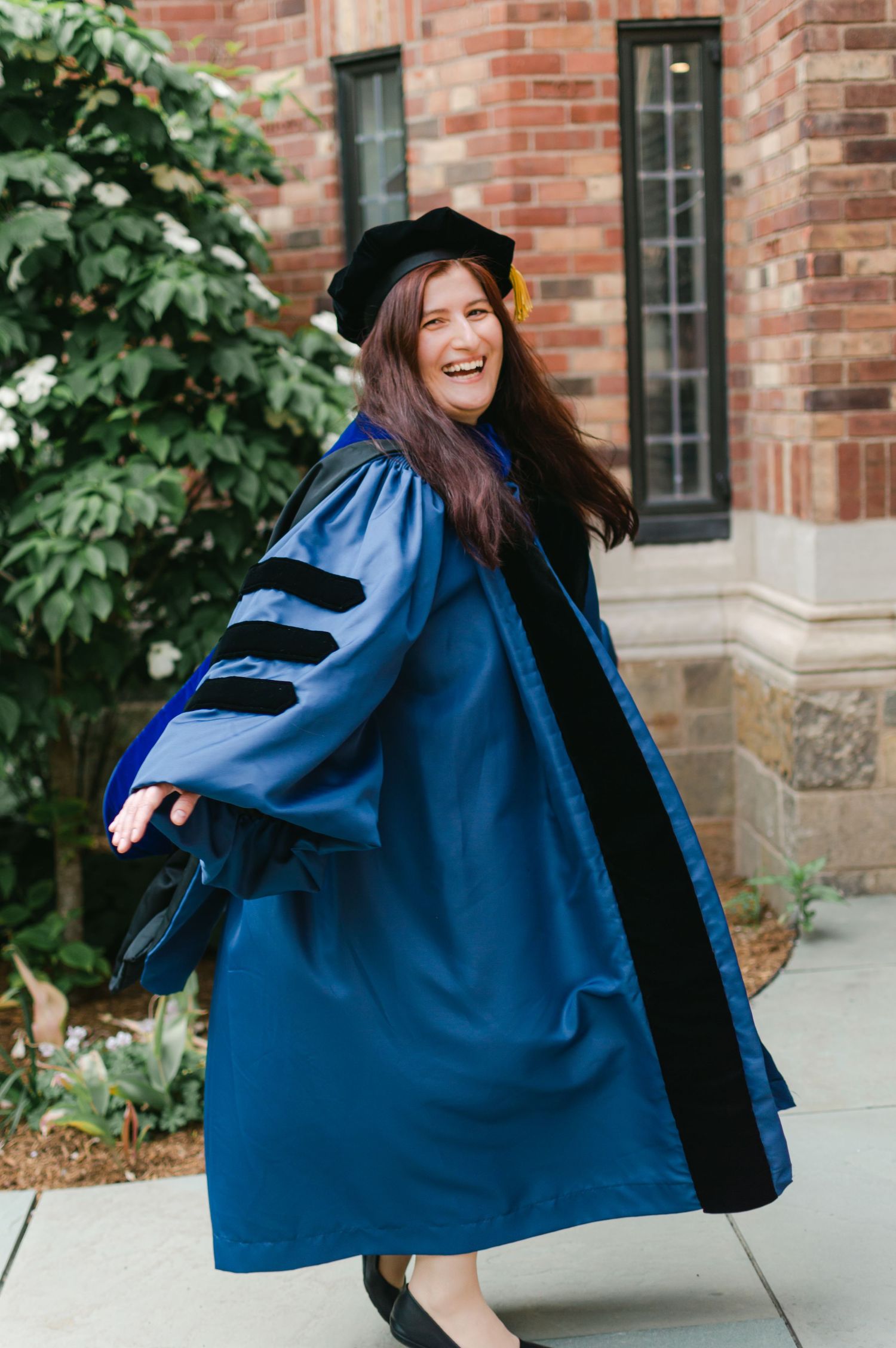 Joyful woman twirling in blue doctoral graduation gown outside brick building.