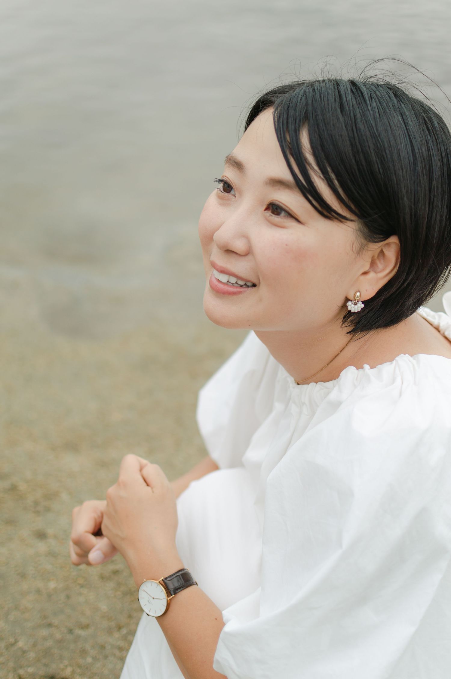 A smiling woman with short hair and floral earrings sits relaxed on a sandy beach shore.