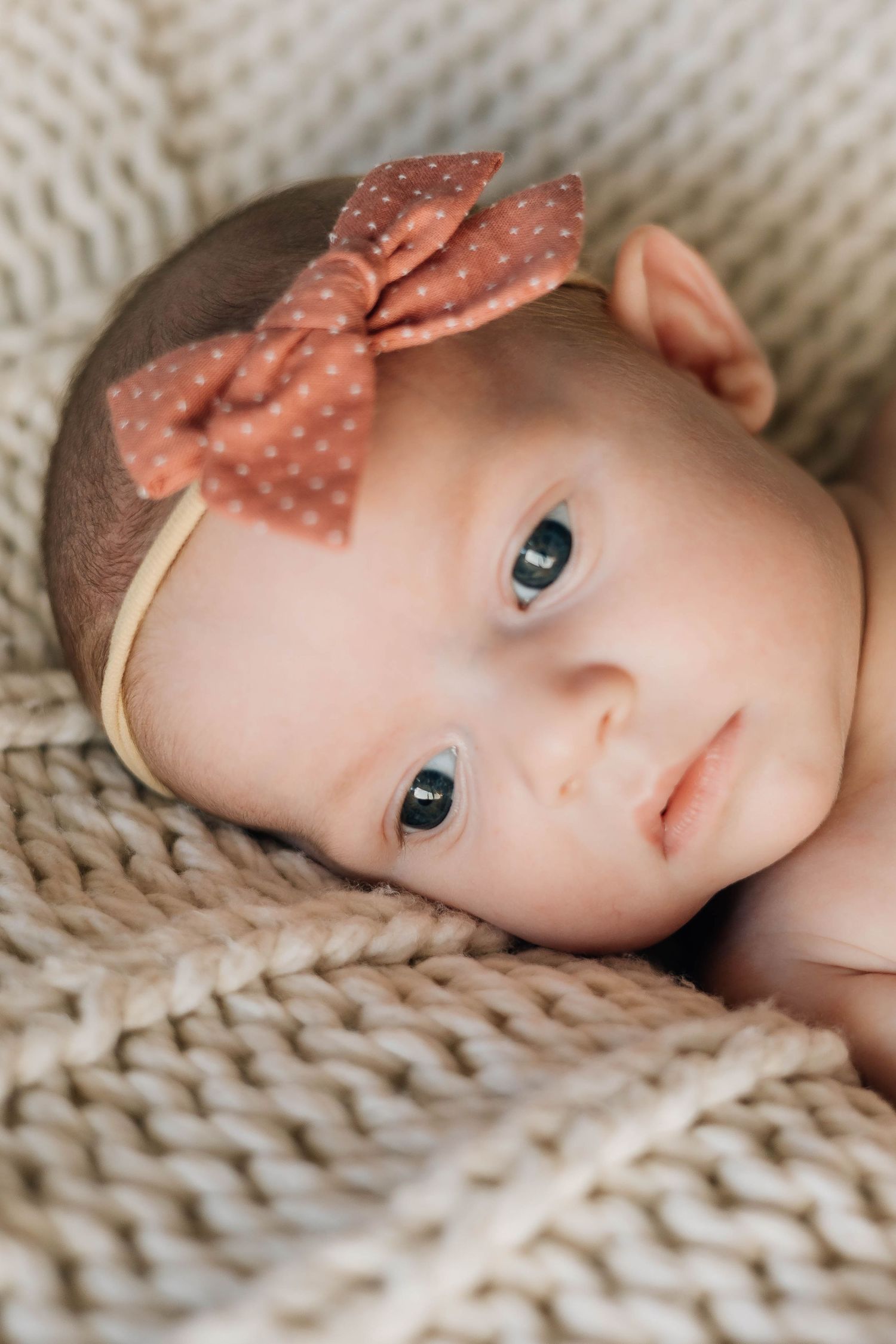 Newborn baby girl with blue eyes wearing a pink polka dot bow headband lying on a knit blanket.