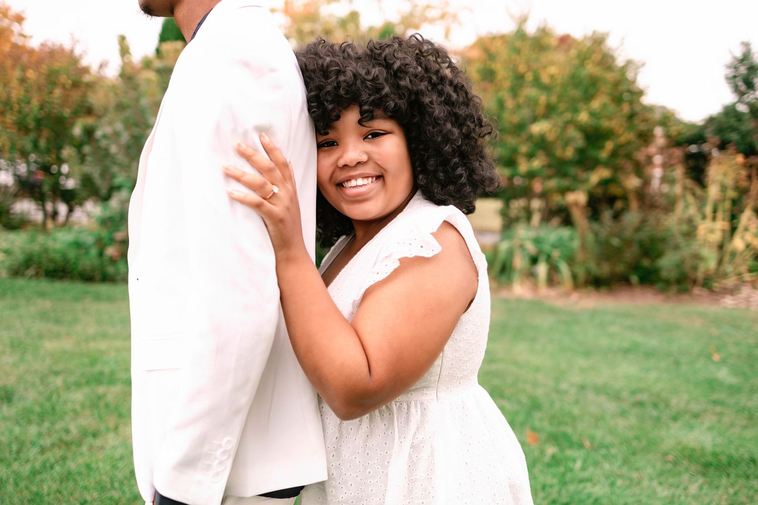 Joyful Black woman with curly hair hugging her partner and smiling at the camera outdoors.