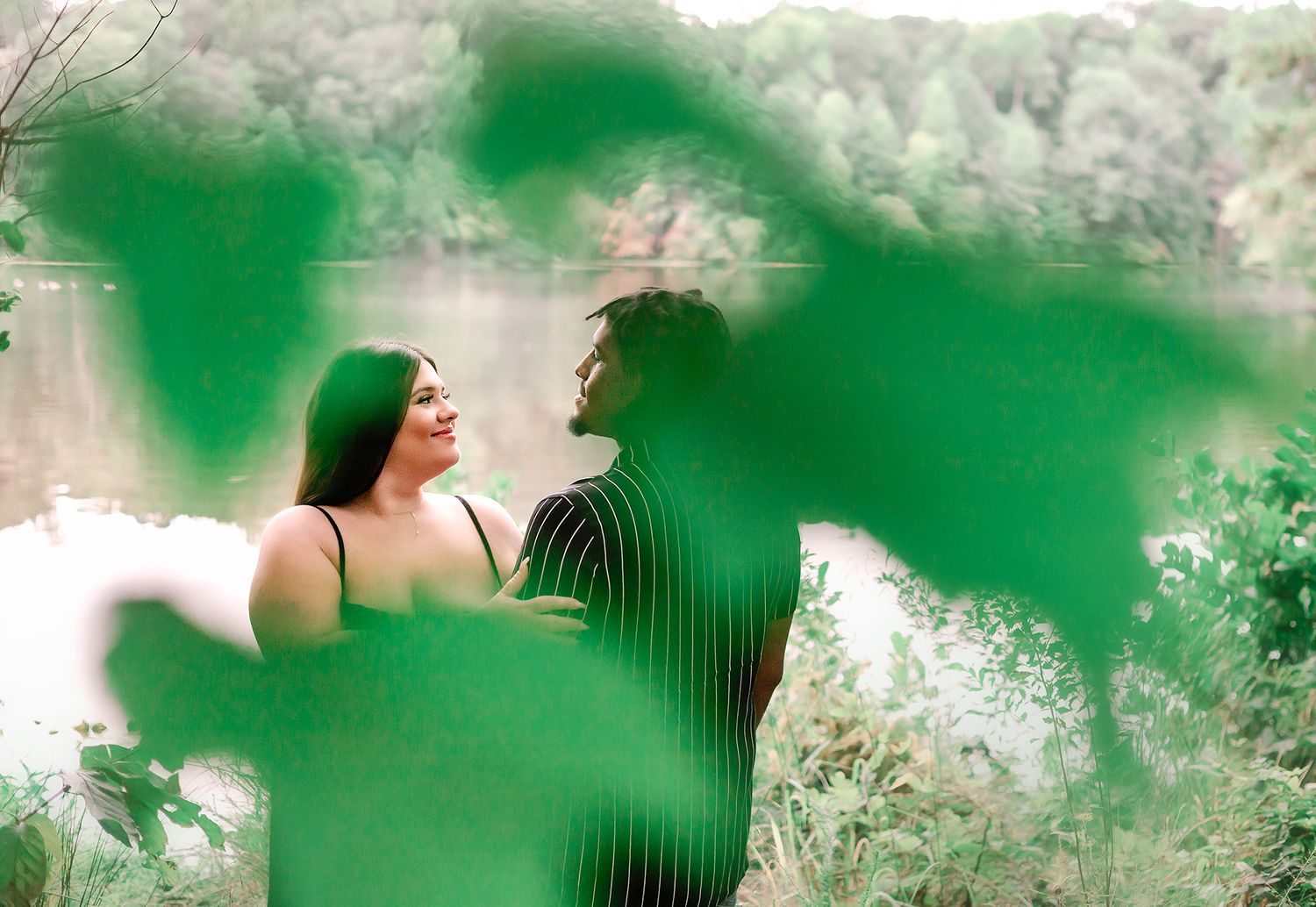 Artistic maternity photo of couple gazing at each other through green leaves by a peaceful lakeside.