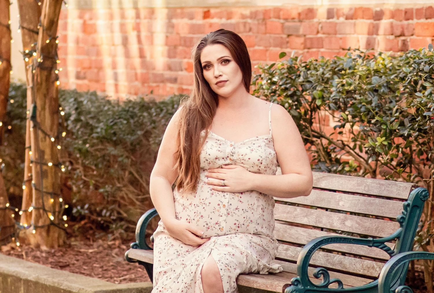 Pregnant woman in floral sundress sitting gracefully on garden bench beside brick wall outdoors.