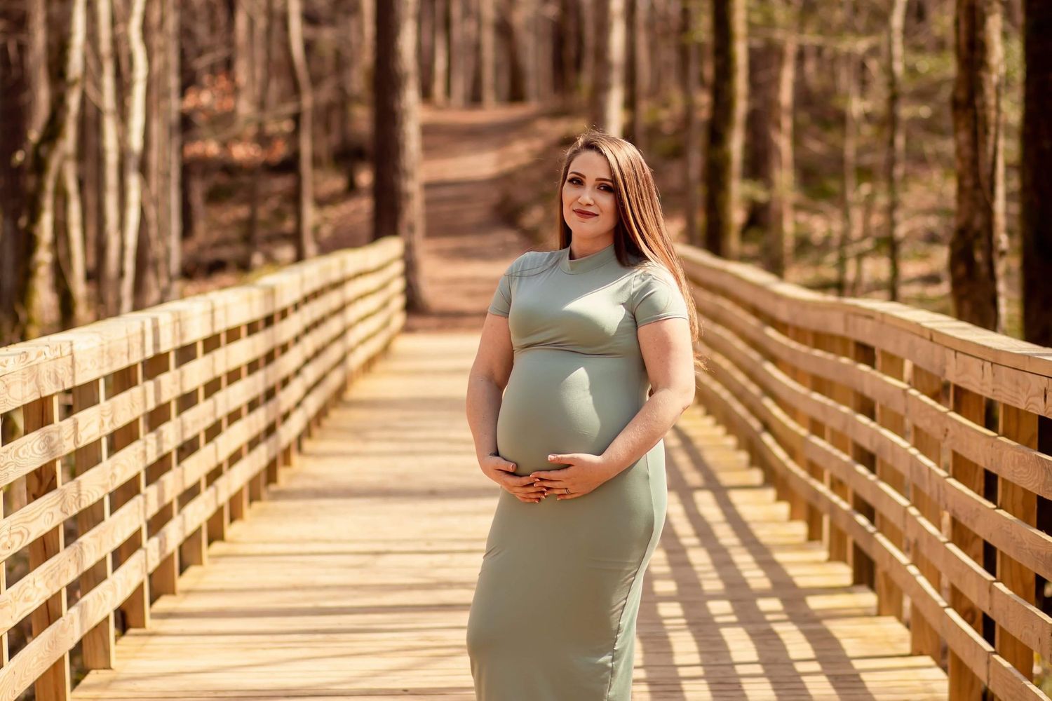 Elegant pregnant woman in sage green dress standing on sunny wooden bridge surrounded by forest.