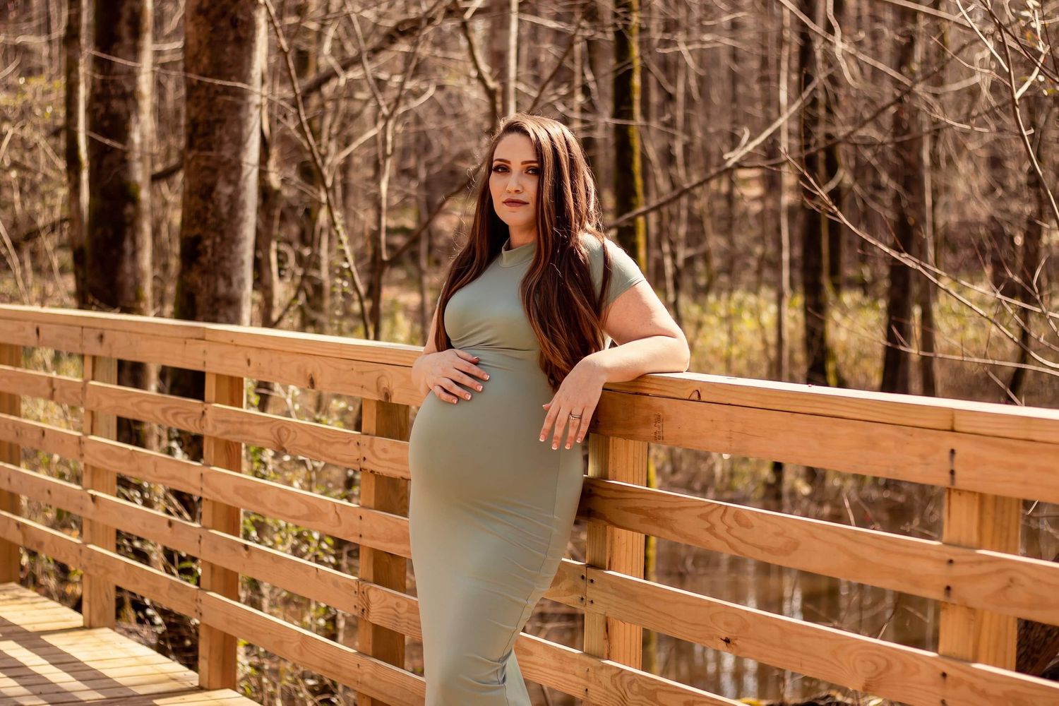 Pregnant woman in fitted sage dress leaning against wooden bridge railing in autumn forest setting.