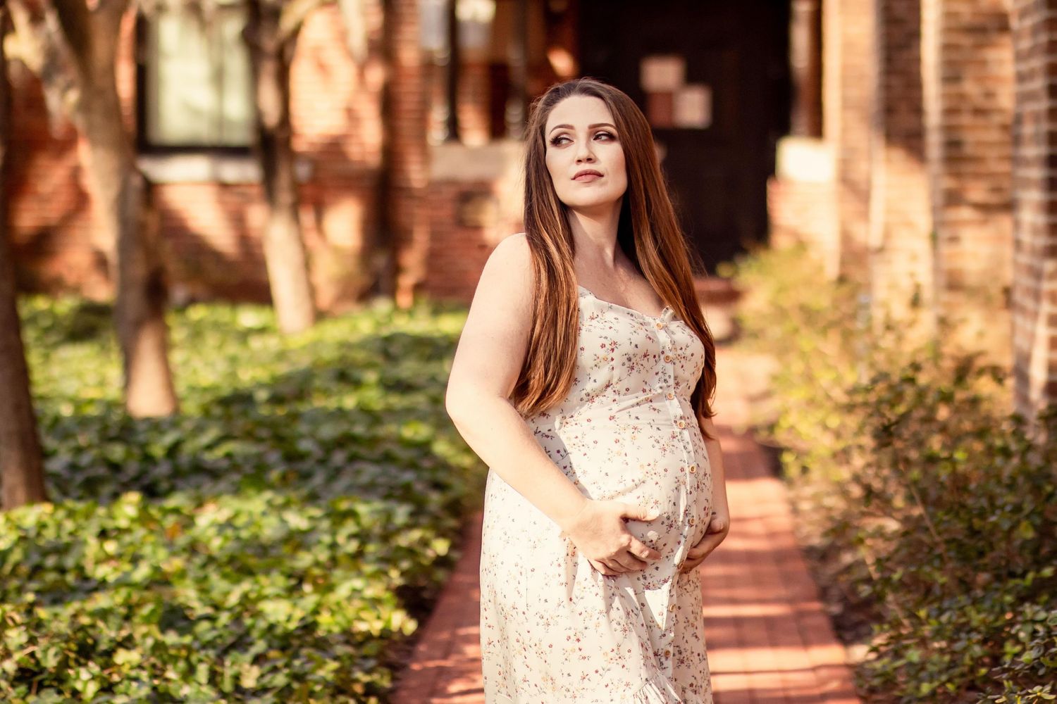 Glowing pregnant woman in floral dress cradling baby bump while standing in sunlit garden path.