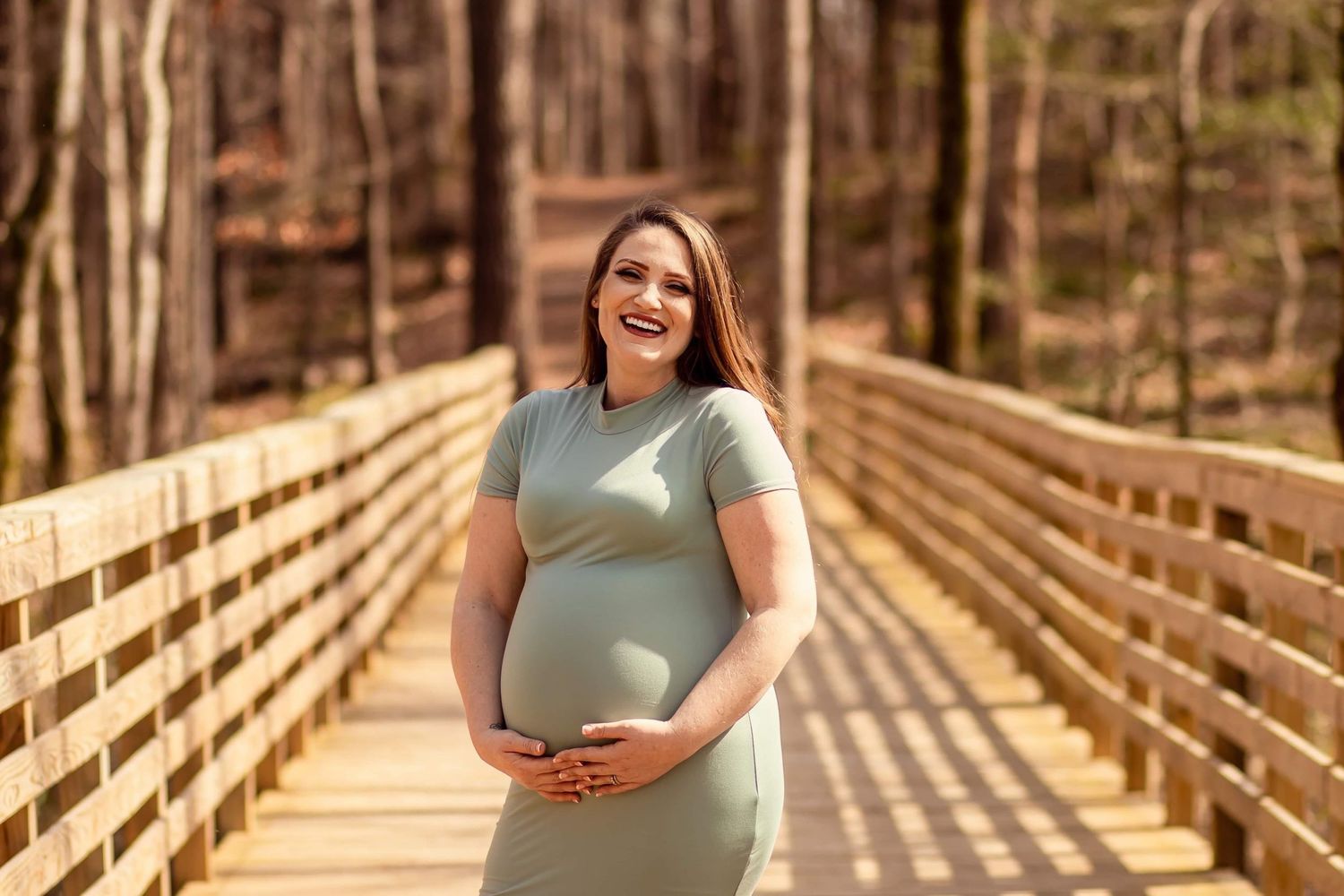 Smiling pregnant woman in sage green dress holding belly on wooden bridge in forest.