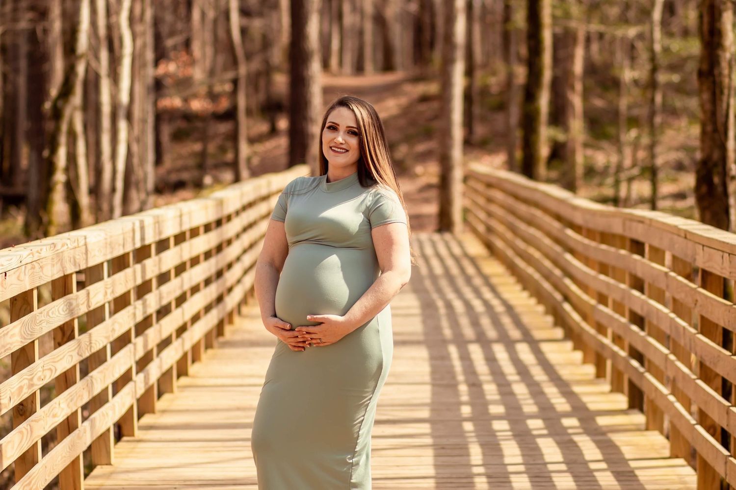 Pregnant woman in sage green dress cradling baby bump on sunlit wooden bridge.